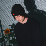 Person wearing a black beanie with an orange emblem, standing against a wire fence with a sunflower in the foreground.