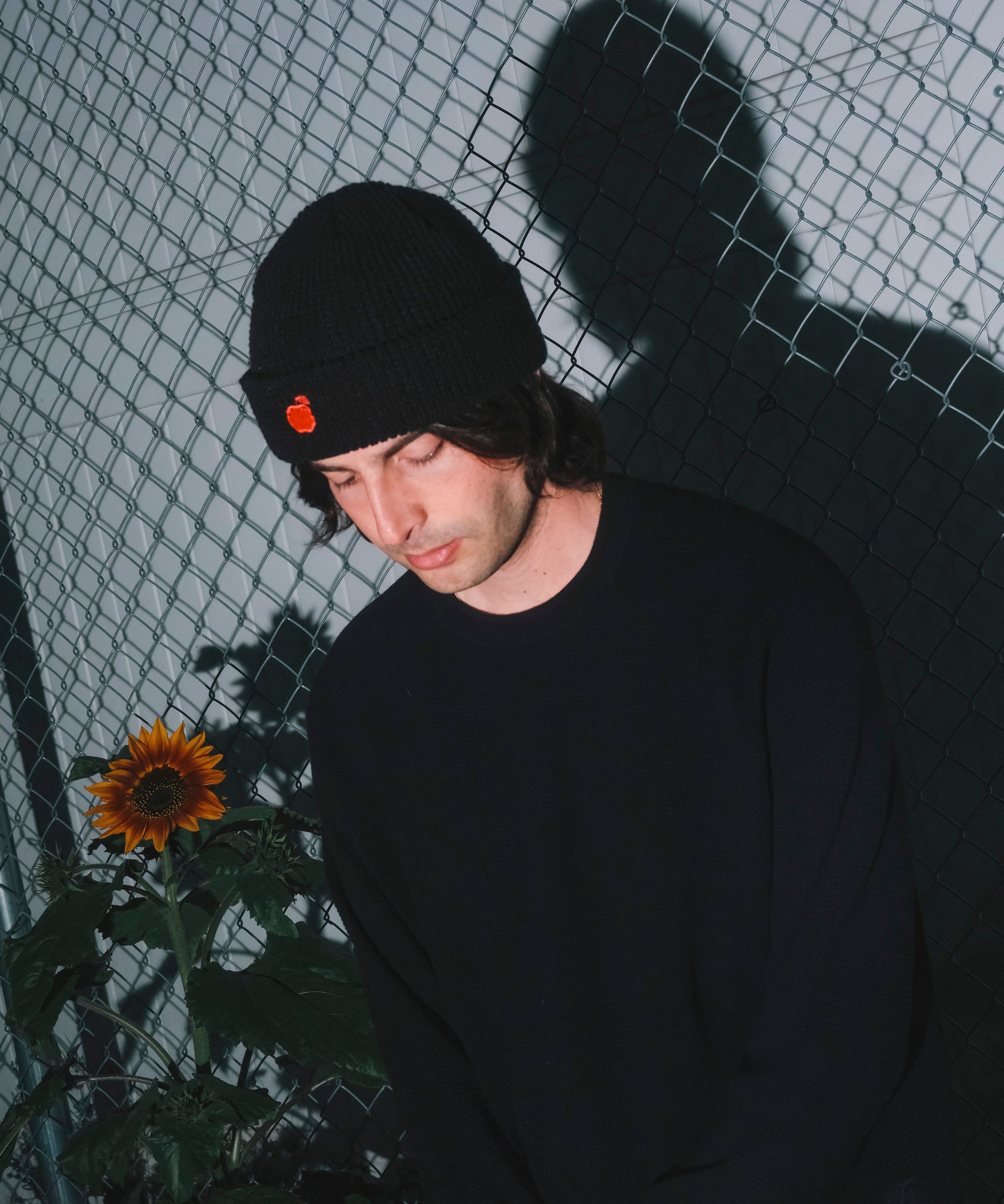 Person wearing a black beanie with an orange emblem, standing against a wire fence with a sunflower in the foreground.