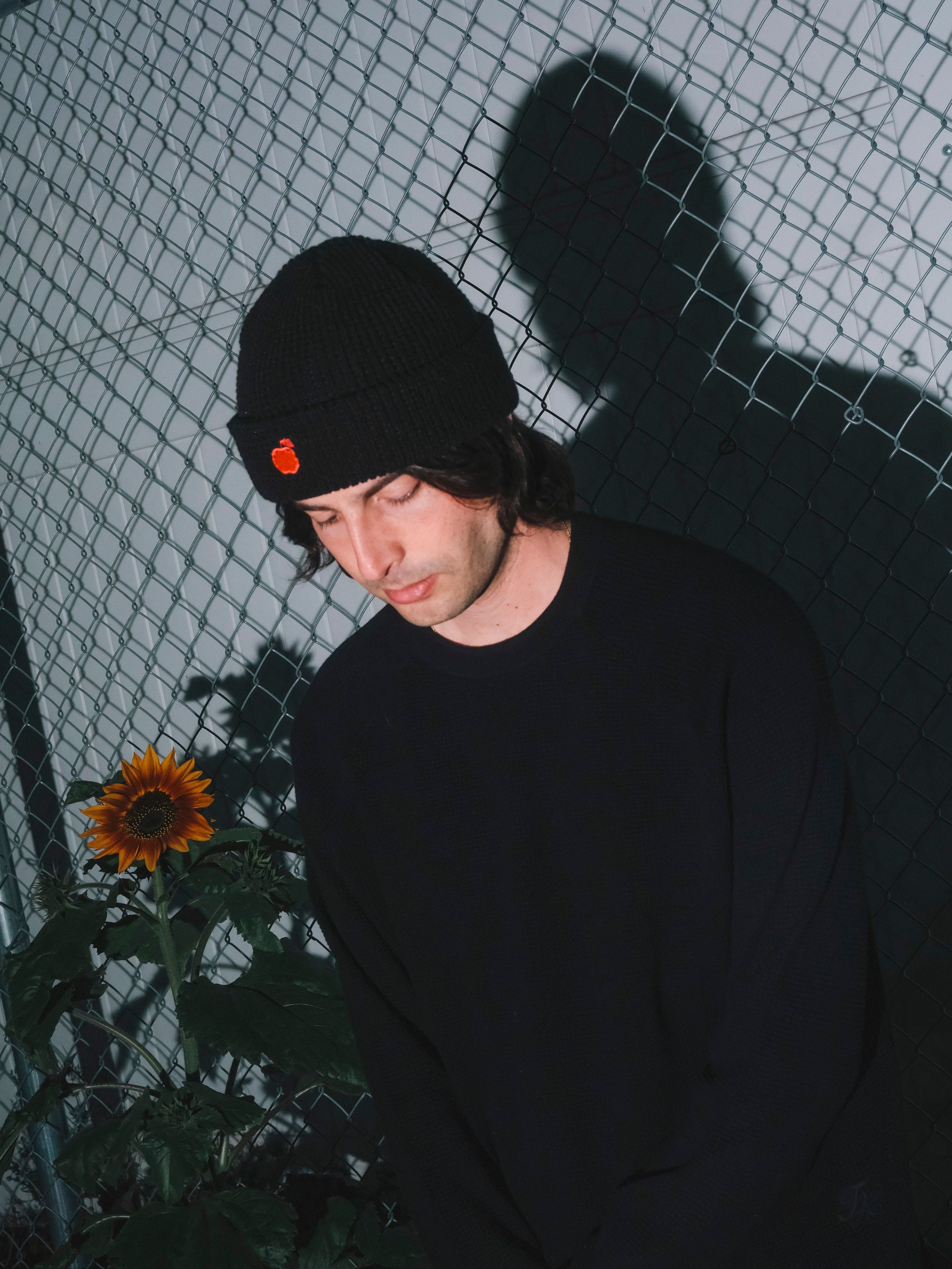 Person wearing a black beanie with an orange emblem, standing against a wire fence with a sunflower in the foreground.