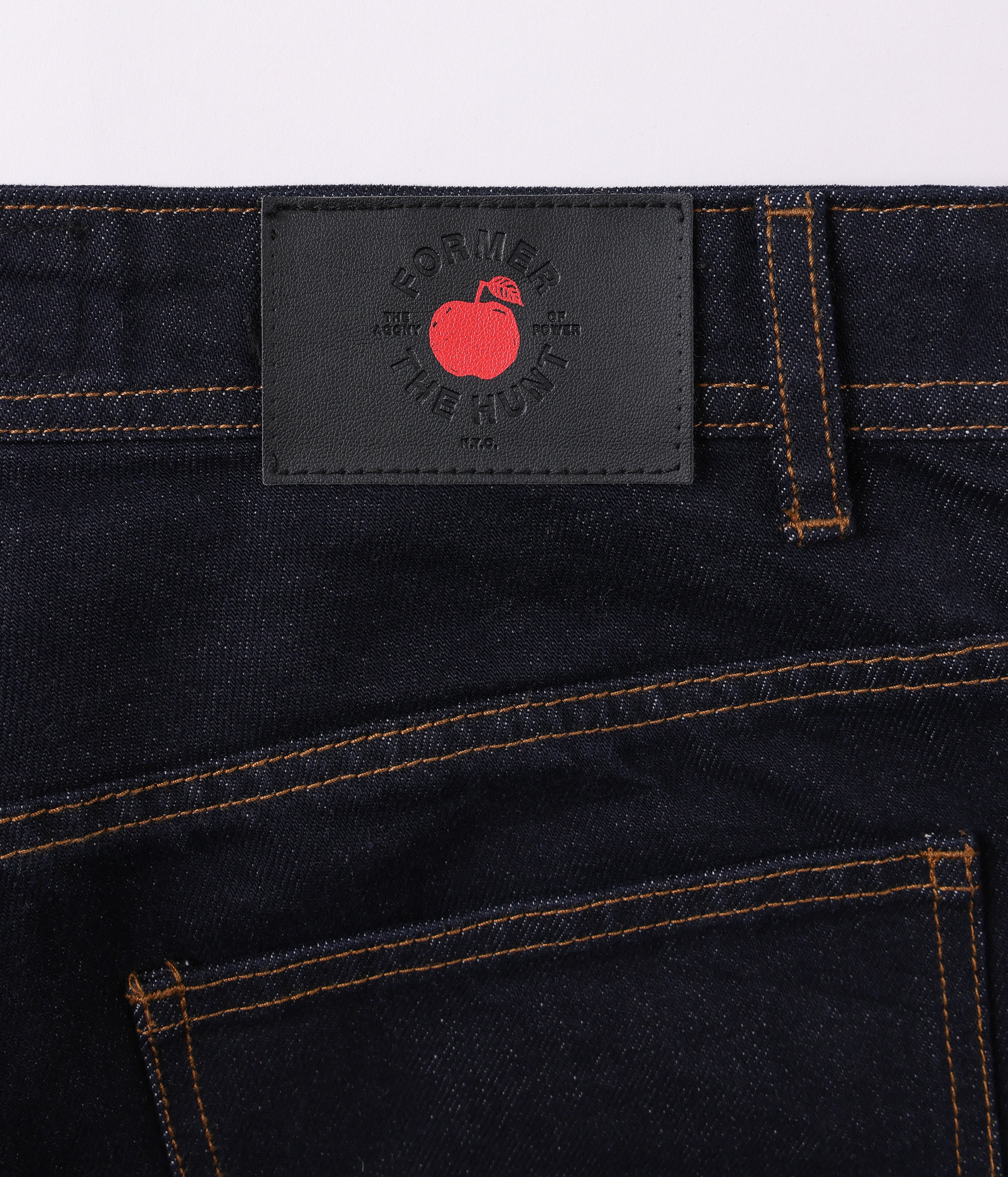 Close-up of dark denim jeans with a brand label featuring a red apple logo on a light gray background.