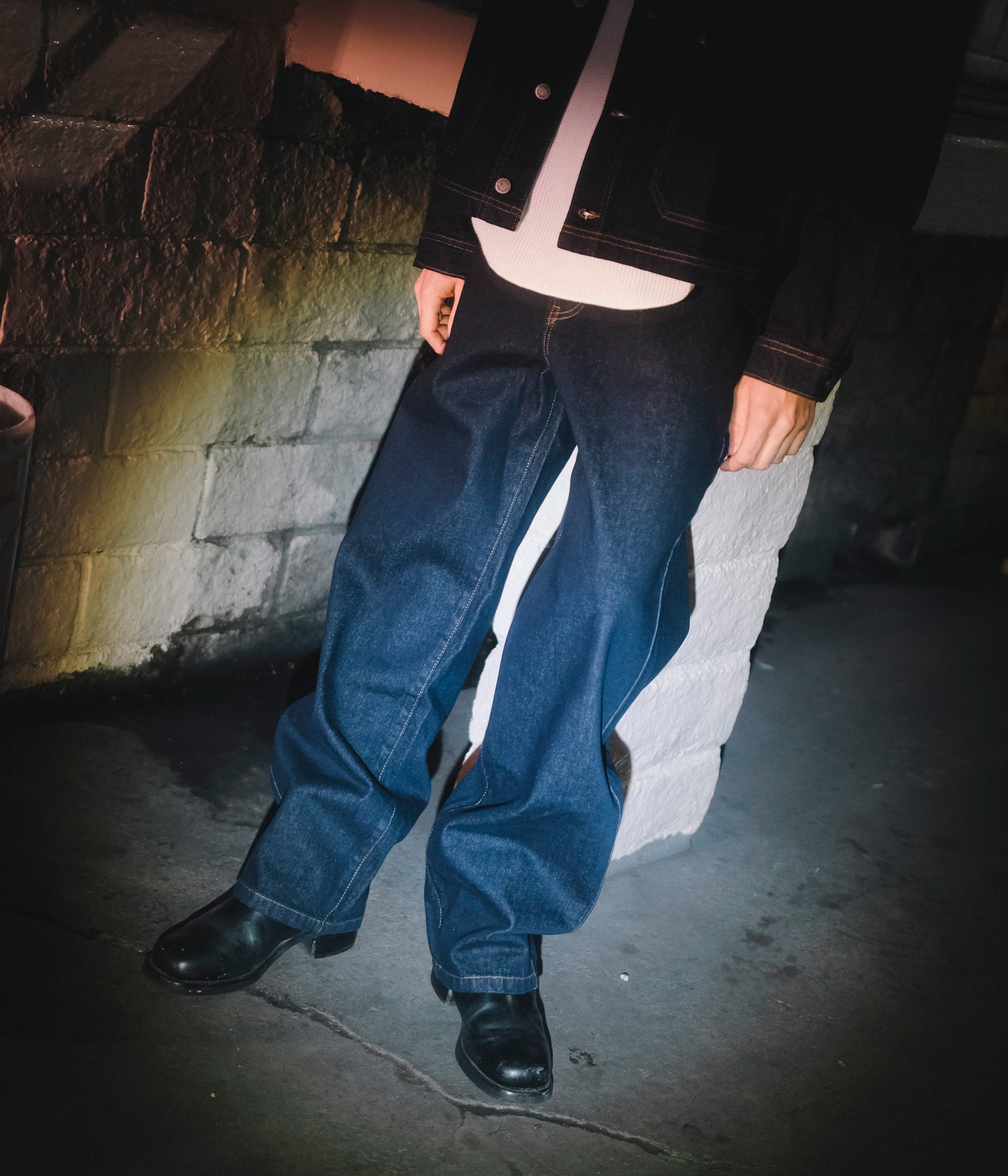 Person wearing a black jacket and blue jeans standing against a stone wall.