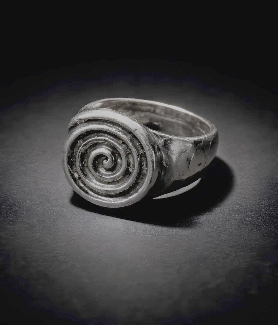 A silver spiral signet ring with a textured, hammered finish, displayed on a dark, reflective surface under dramatic lighting.