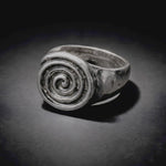 A silver spiral signet ring with a textured, hammered finish, displayed on a dark, reflective surface under dramatic lighting.