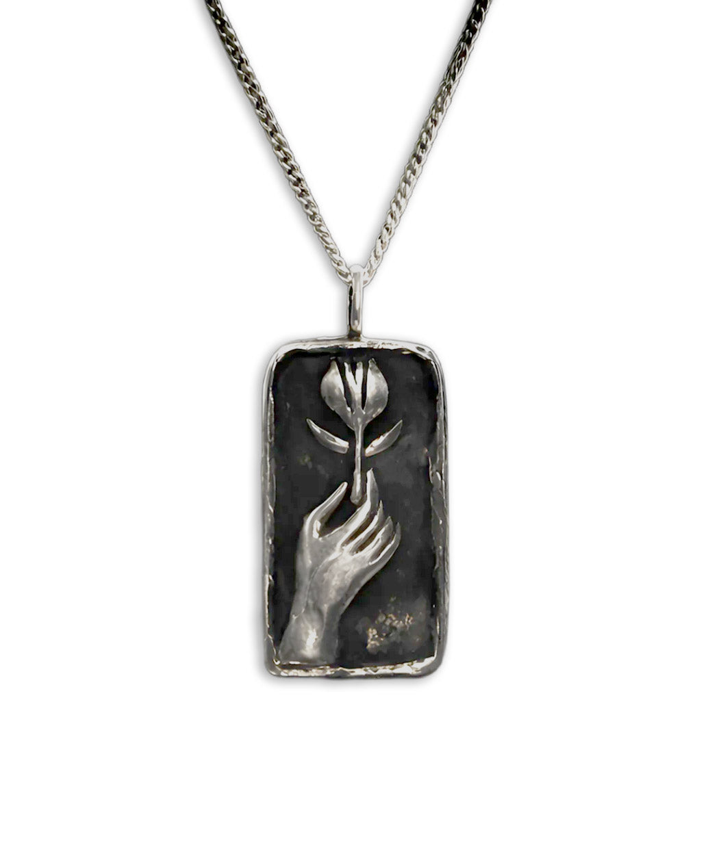 A rectangular silver pendant necklace featuring an embossed hand holding a tulip, suspended from a silver curb chain against a plain white background.