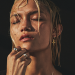 Close-up of a woman with closed eyes, wearing gold earrings and rings, against a dark background.
