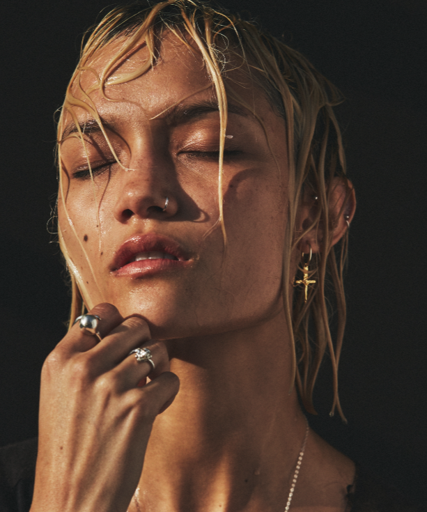 Close-up of a woman with closed eyes, wearing gold earrings and rings, against a dark background.
