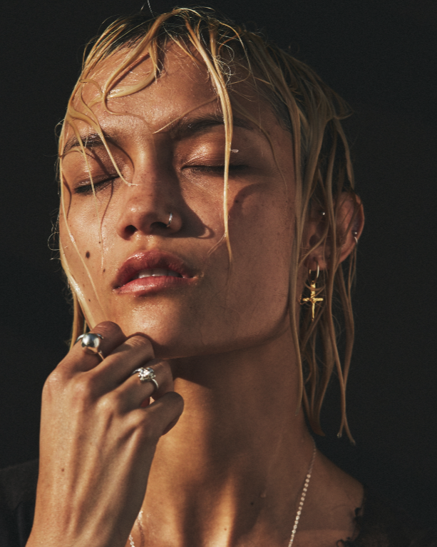 Close-up of a woman with closed eyes, wearing gold earrings and rings, against a dark background.