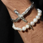 Silver bracelet with a cross design and pearl bracelet on a wrist against a dark background