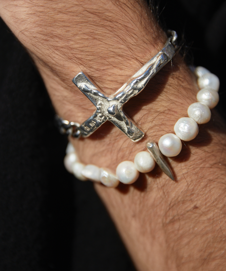 Silver bracelet with a cross design and pearl bracelet on a wrist against a dark background