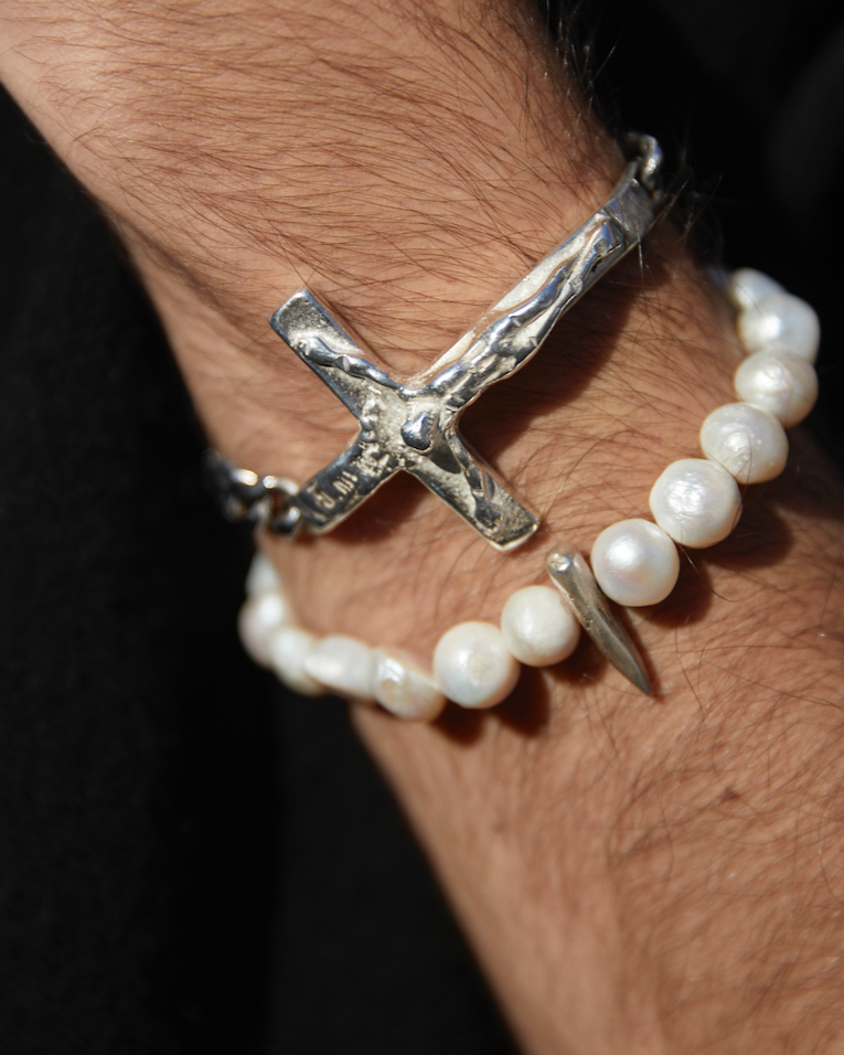 Silver bracelet with a cross design and pearl bracelet on a wrist against a dark background