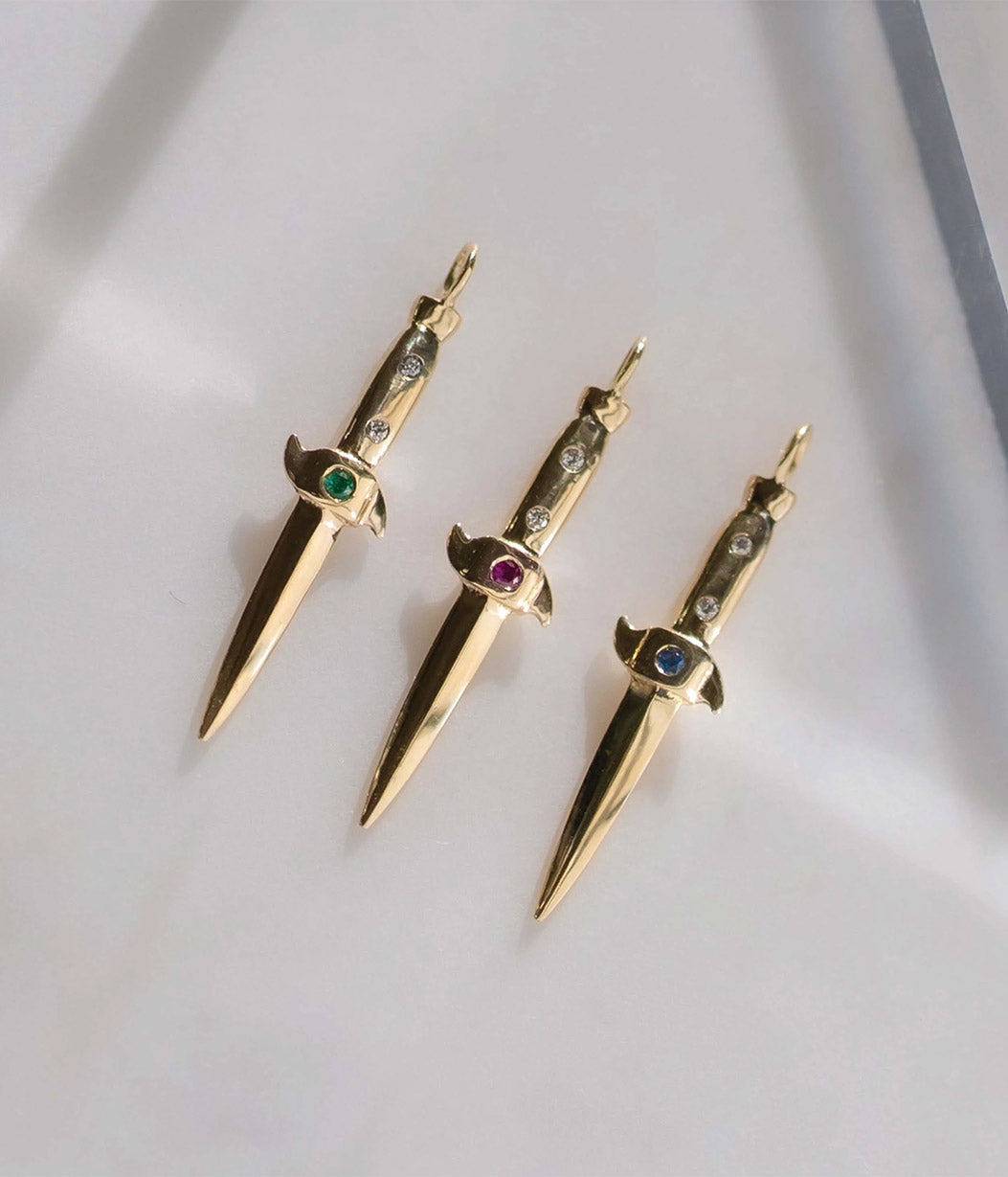 Three gold dagger-shaped pendants with gemstones on a light gray background