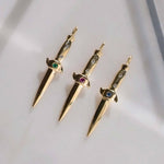 Three gold dagger-shaped pendants with gemstones on a light gray background