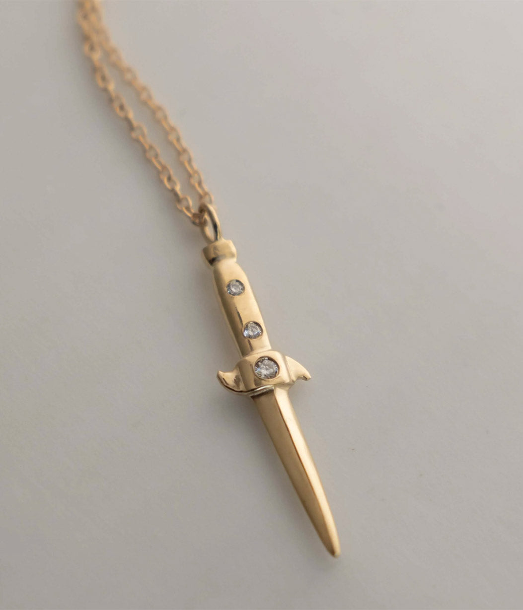 Gold dagger-shaped necklace with embedded stones on a light gray background