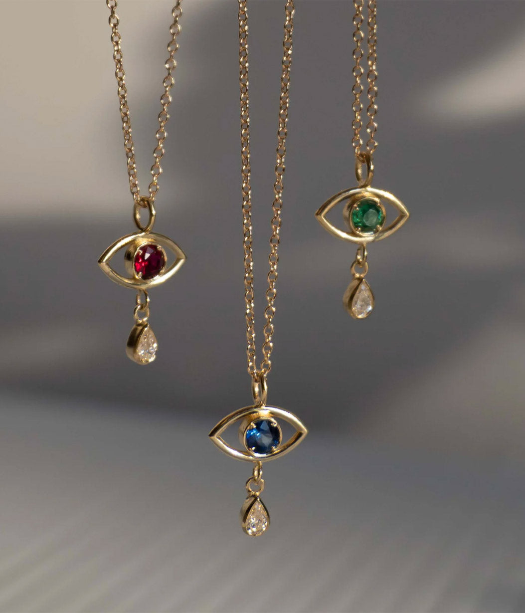Three gold necklaces with eye-shaped pendants featuring gemstones on a gray background.