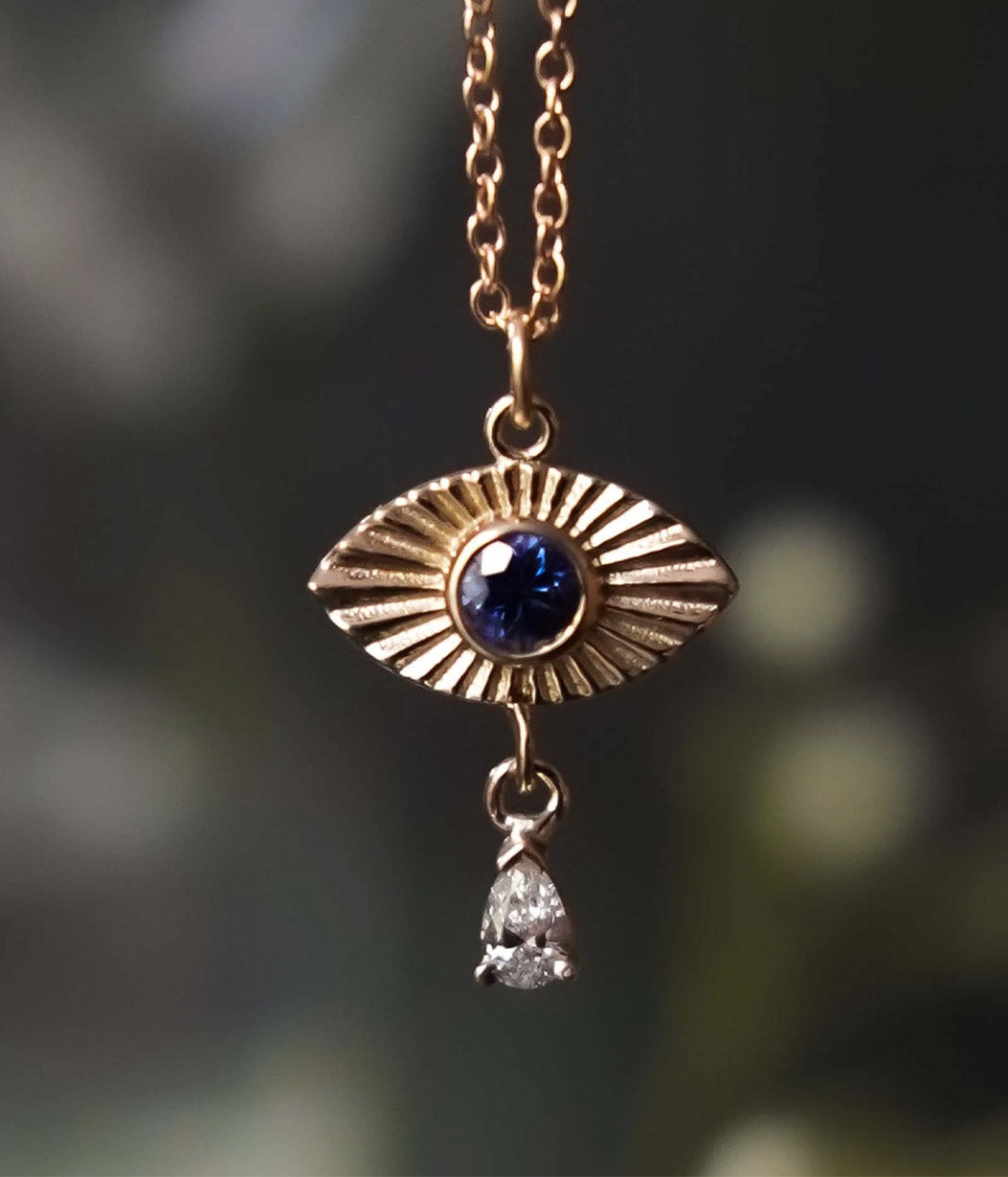 Gold pendant with blue gemstone and diamond drop against a blurred natural background
