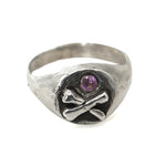 Silver ring with skull and crossbones design and purple gemstone on a white background