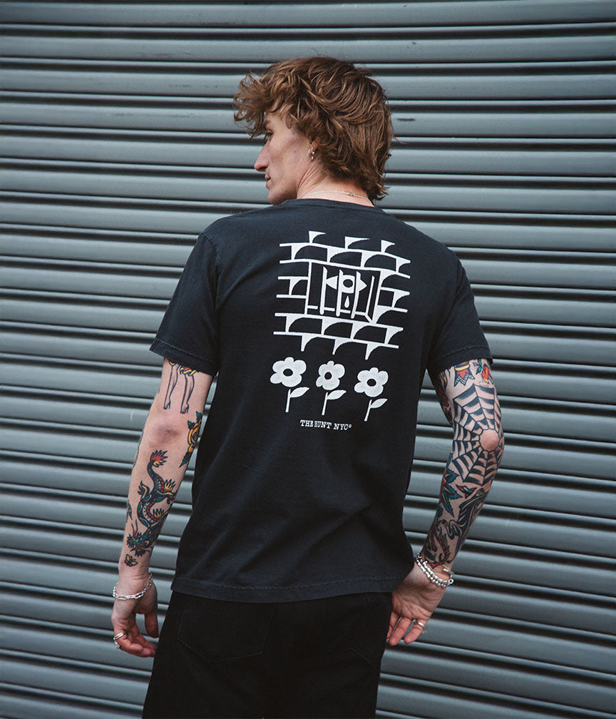 Person wearing a black t-shirt with white graphics and text, standing in front of a metallic shutter.