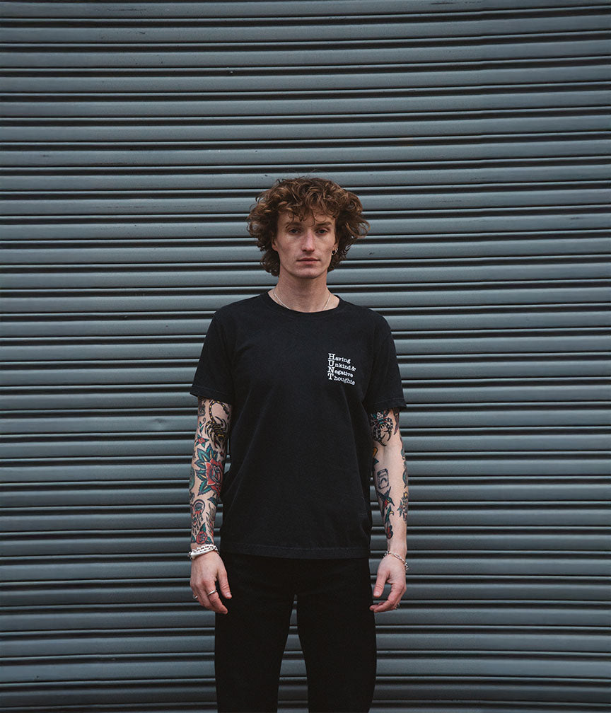 Person wearing a black t-shirt with a logo in front of a gray metal shutter.