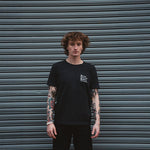 Person wearing a black t-shirt with a logo in front of a gray metal shutter.