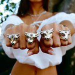 Four gold rings with bull head designs on a person's fingers outdoors.