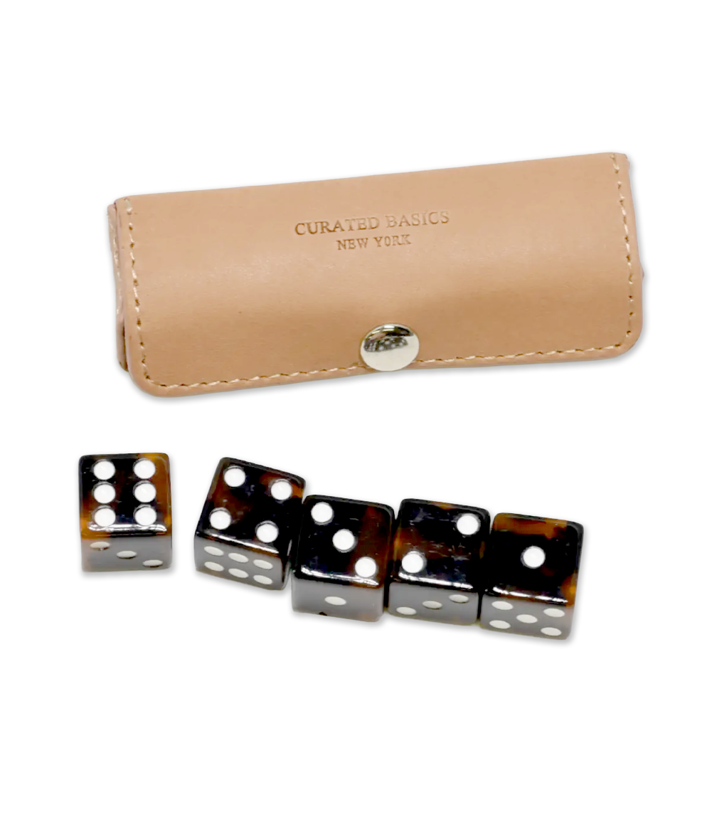 A set of five black and brown tortoiseshell dice with white pips, shown with a beige leather case embossed with 'CURATED BASICS NEW YORK' and a silver snap closure, on a white background.