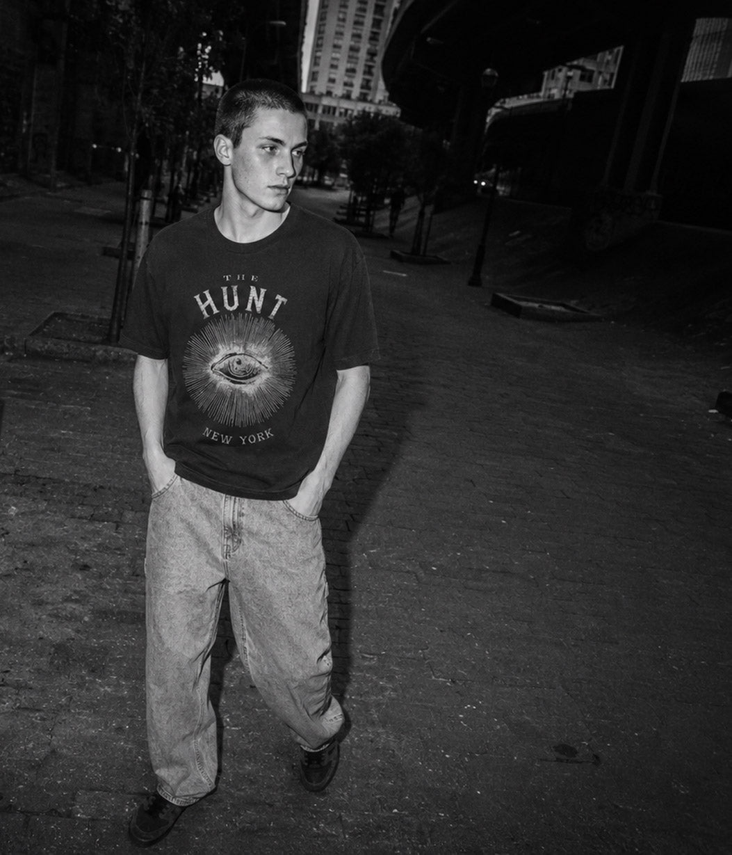 A man standing outdoors at night wearing a dark graphic T-shirt with 'THE HUNT NEW YORK' and an eye design, paired with light-colored jeans.