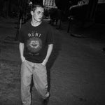 A man standing outdoors at night wearing a dark graphic T-shirt with 'THE HUNT NEW YORK' and an eye design, paired with light-colored jeans.