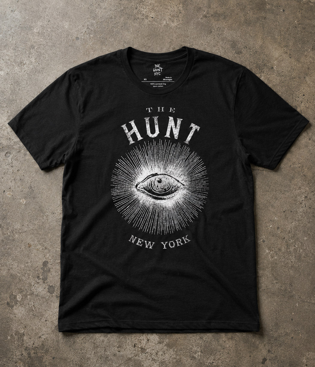 A black short-sleeve graphic t-shirt laid flat on a textured concrete surface. The shirt features a white distressed print of an eye surrounded by radiating lines, with the text 'THE HUNT' above and 'NEW YORK' below.