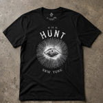 A black short-sleeve graphic t-shirt laid flat on a textured concrete surface. The shirt features a white distressed print of an eye surrounded by radiating lines, with the text 'THE HUNT' above and 'NEW YORK' below.