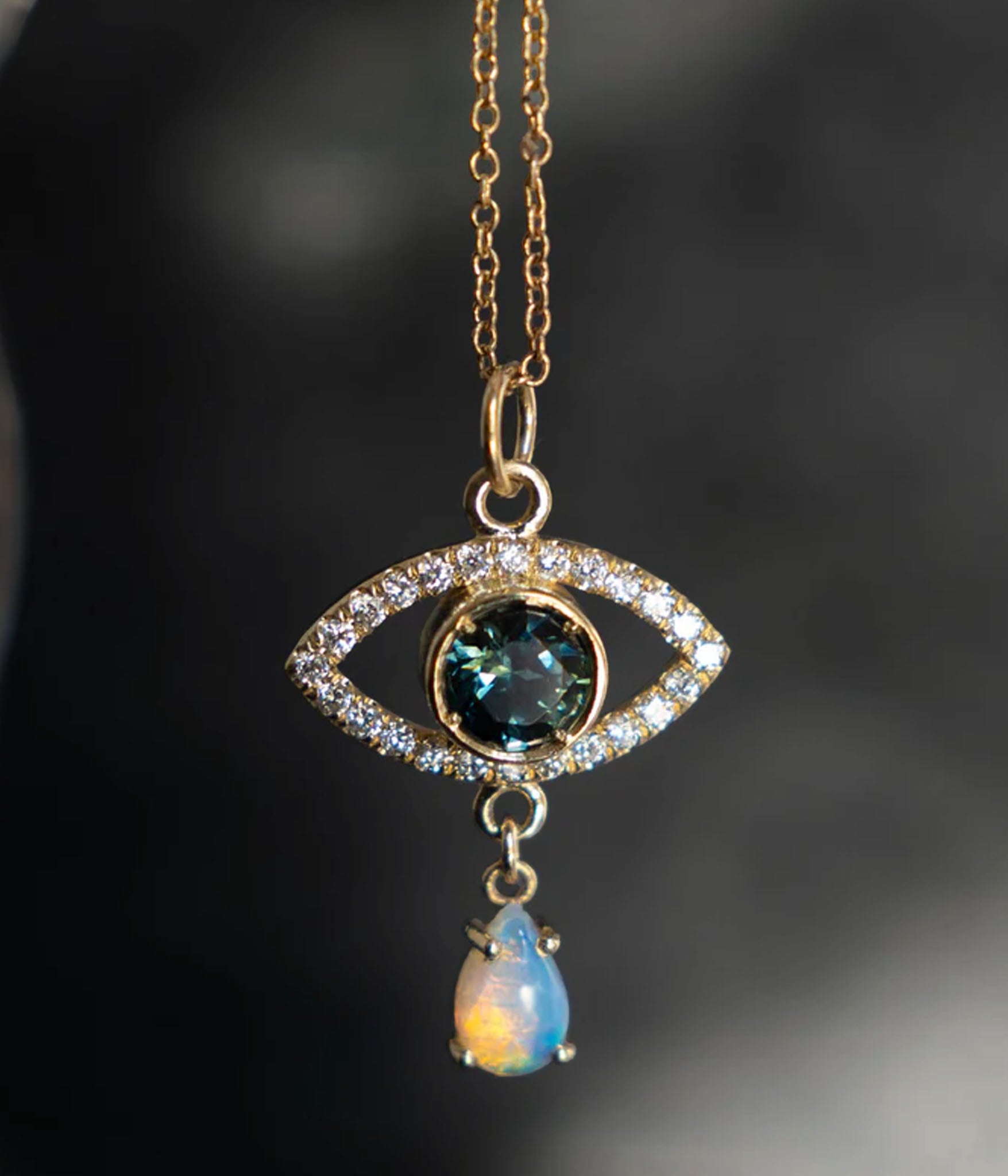 A breathtaking eye amulet featuring a faceted sapphire center stone, surrounded by 14 pavé-set diamonds, and adorned with an opal teardrop.