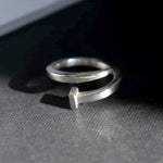 Handmade sterling silver ring shaped like a bent nail, positioned on edge in soft raking light. Surface scratches and subtle patina visible, reinforcing handcrafted quality.