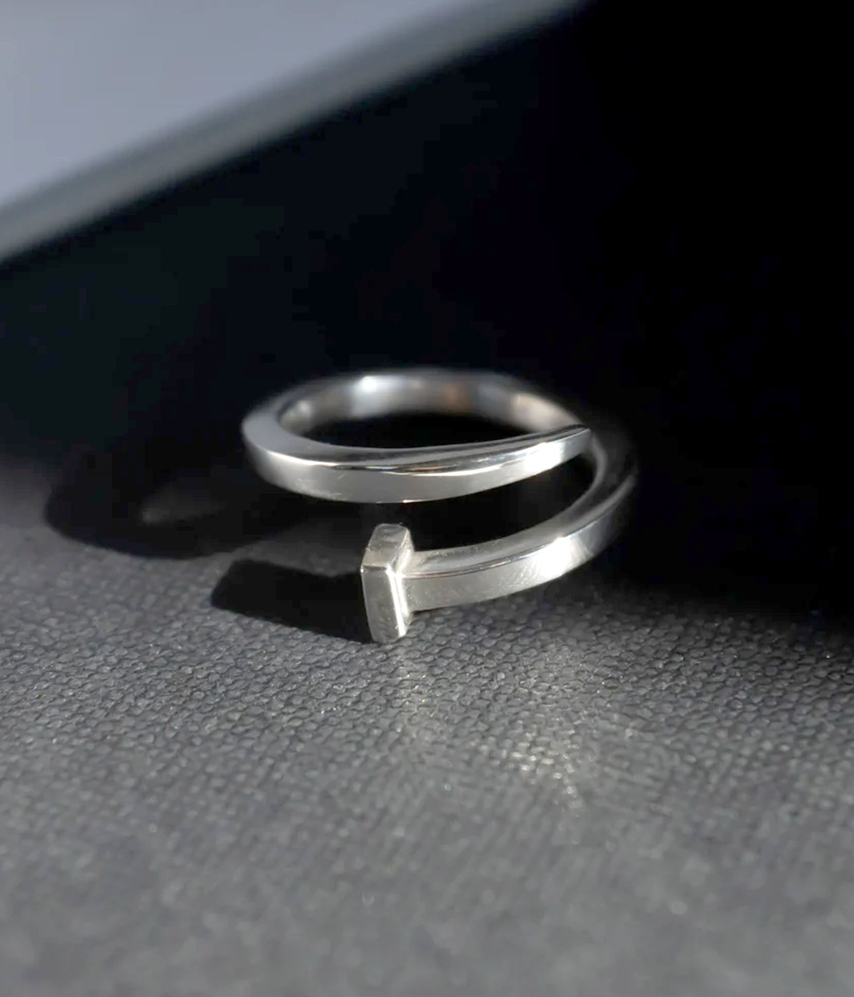 Handmade sterling silver ring shaped like a bent nail, positioned on edge in soft raking light. Surface scratches and subtle patina visible, reinforcing handcrafted quality.