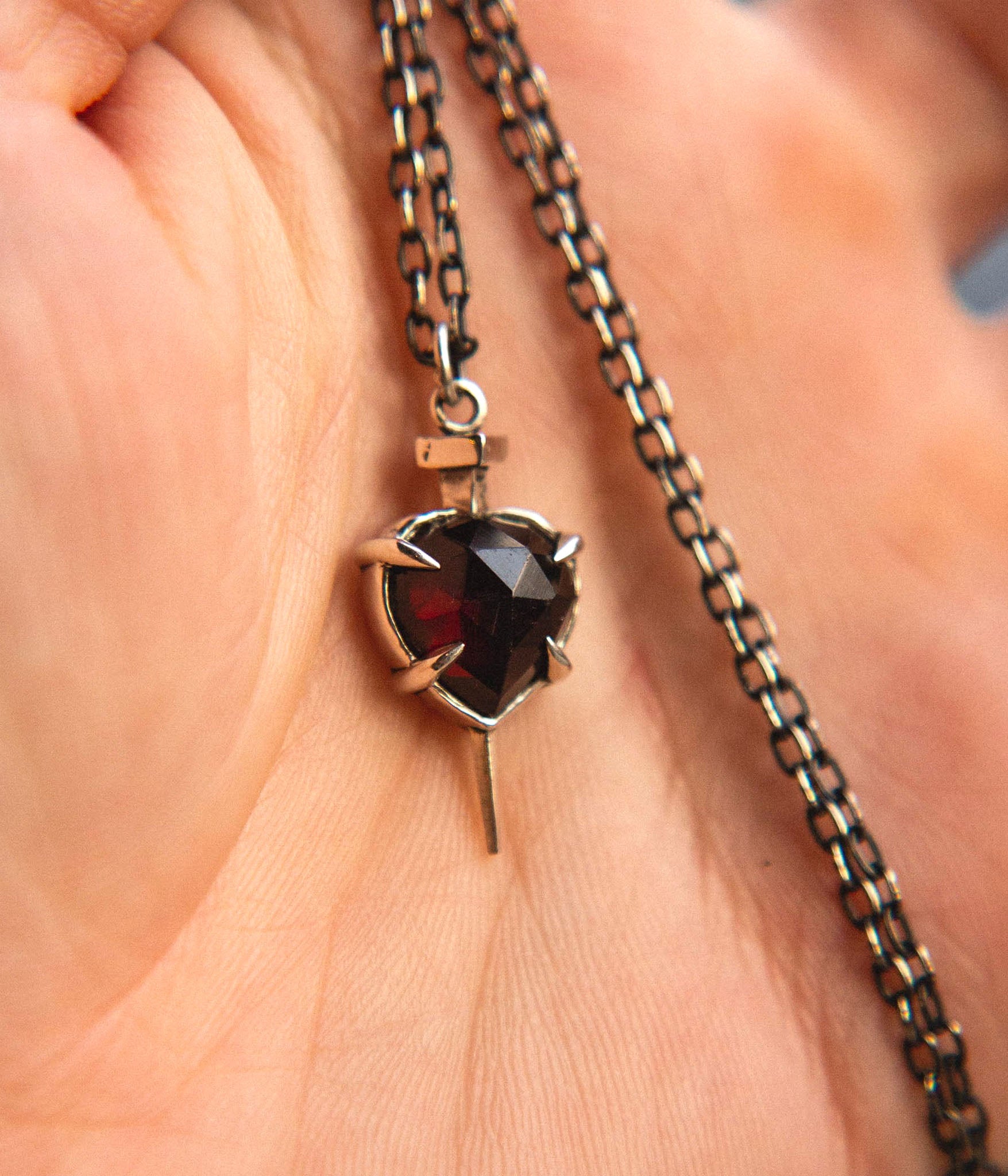 Close-up of hand holding nail-through-heart garnet pendant on oxidized chain