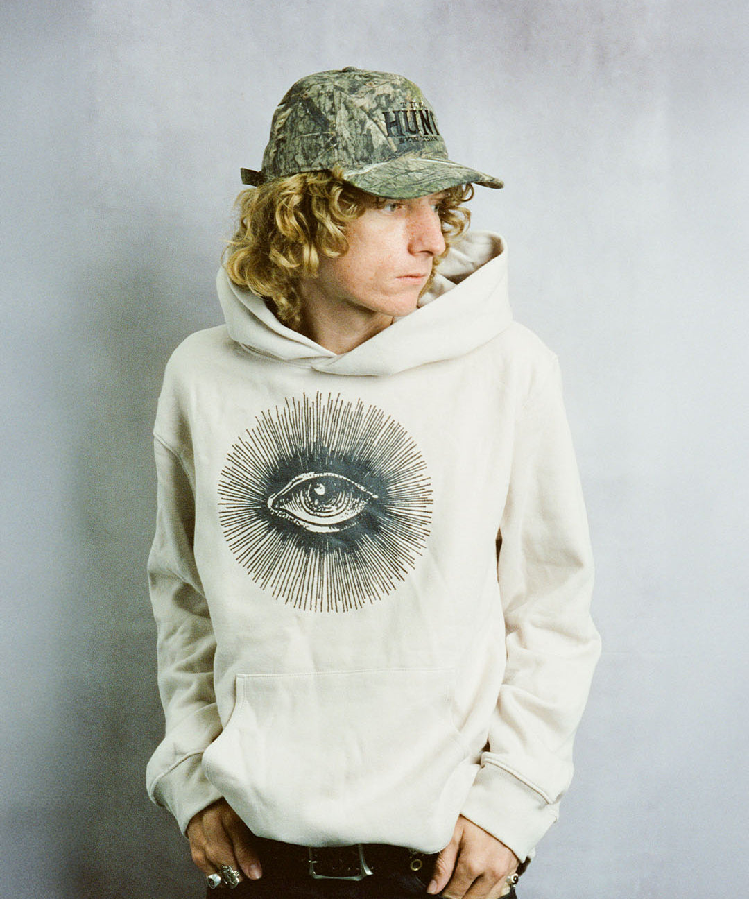 Person wearing a beige hoodie with an eye design and a camouflage cap on a plain background
