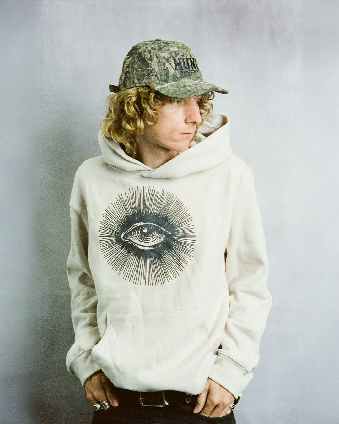 Person wearing a beige hoodie with an eye design and a camouflage cap on a plain background