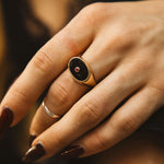 Close-up of a hand wearing a gold ring with a dark stone on a blurred background