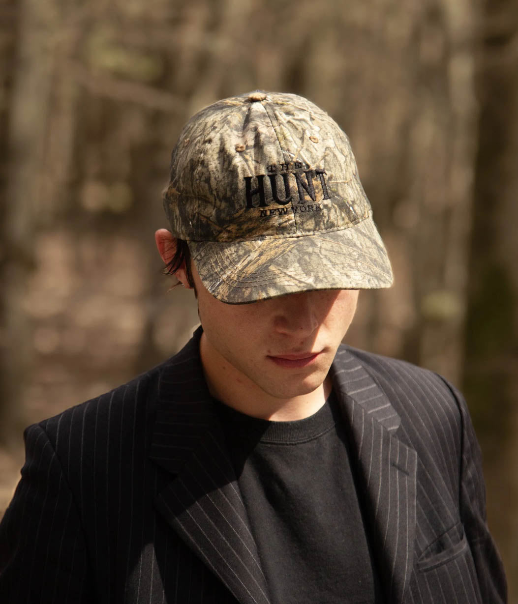 Person wearing a camouflage cap with 'HUNT' printed on it, standing in a forest.