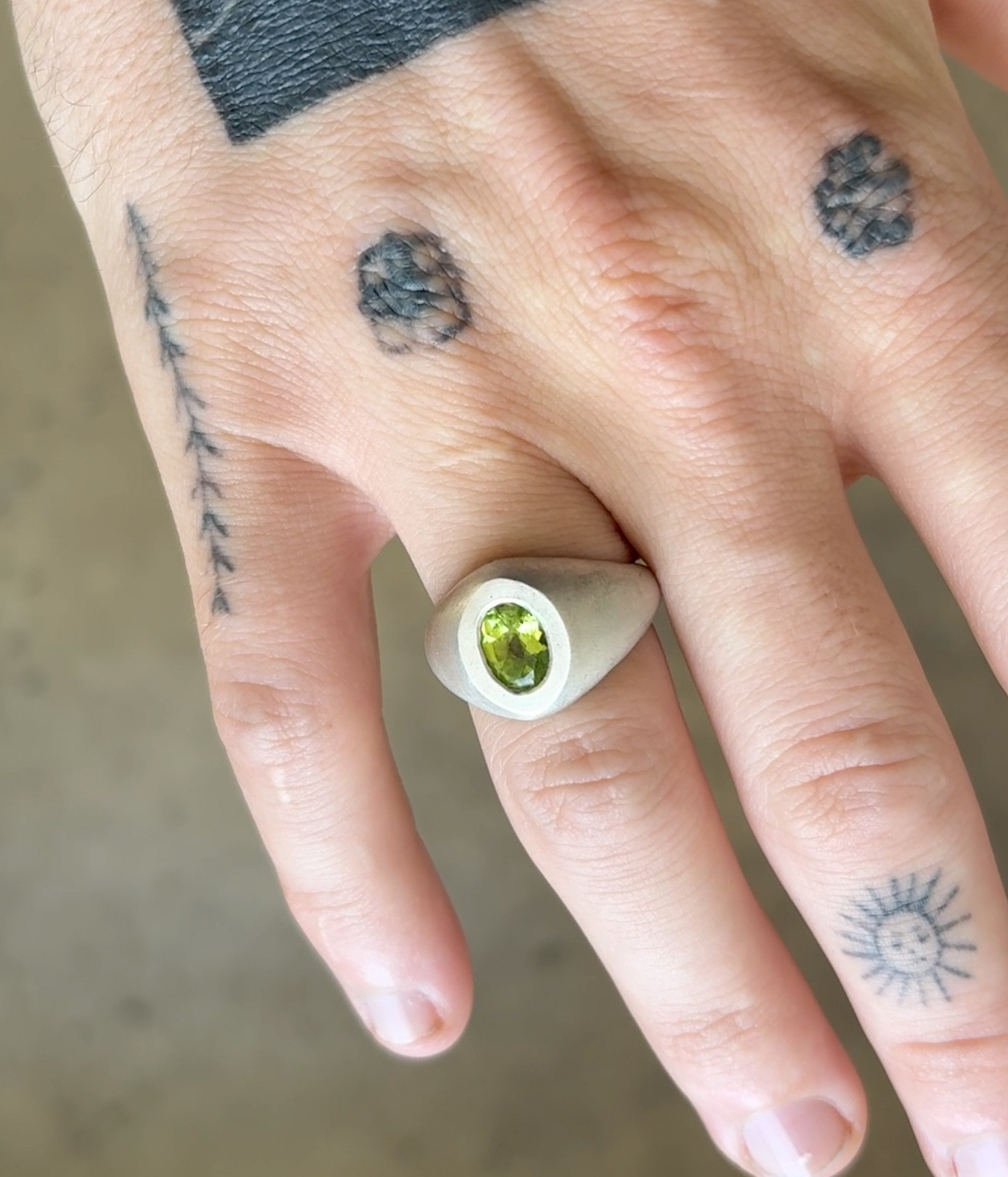 Hand wearing a silver ring with a green gemstone, surrounded by tattoos on a neutral background