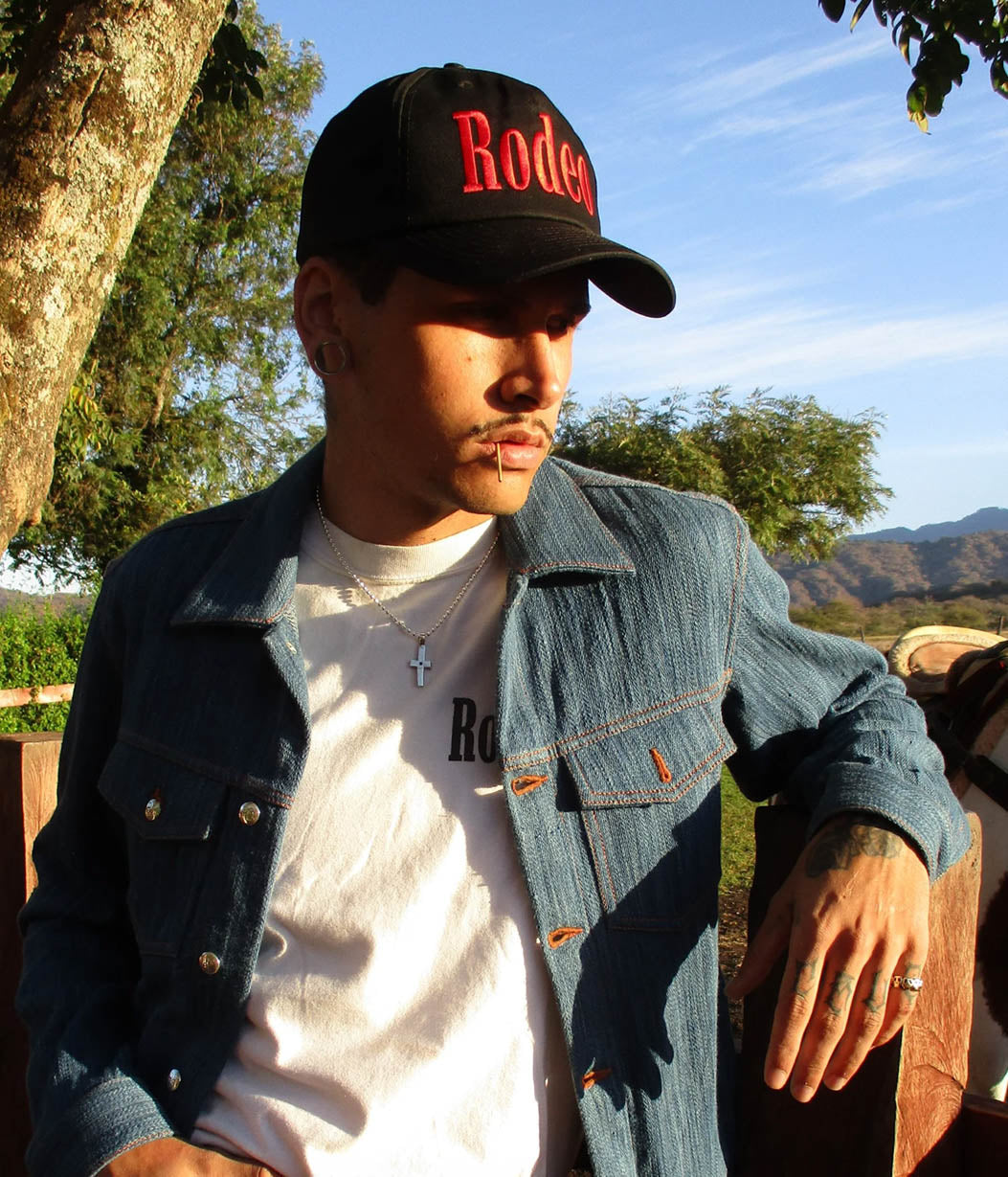 Man wearing a black cap with 'Rodeo' text, denim jacket, and white shirt outdoors.