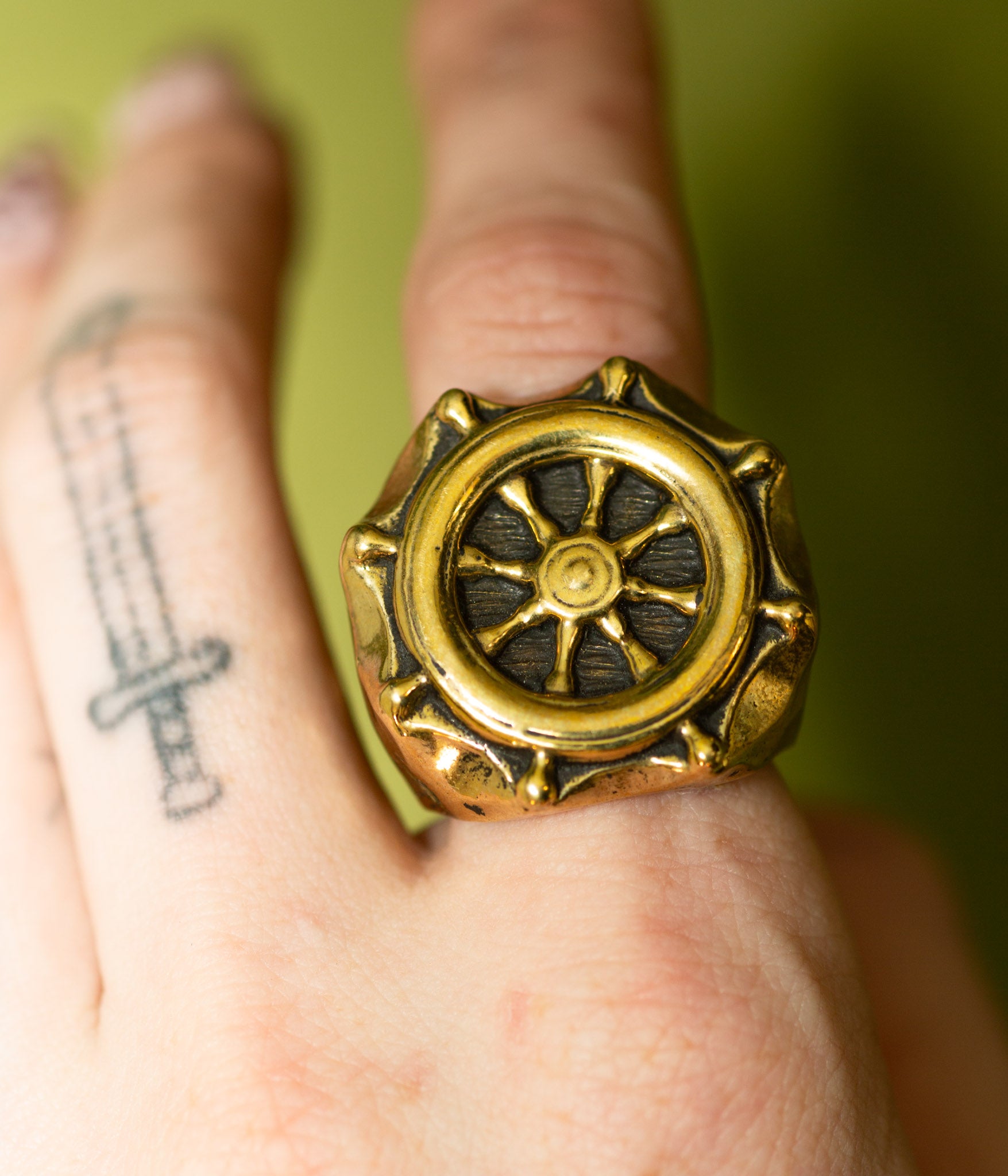 Gold ring with a wheel design on a person's finger against a green background