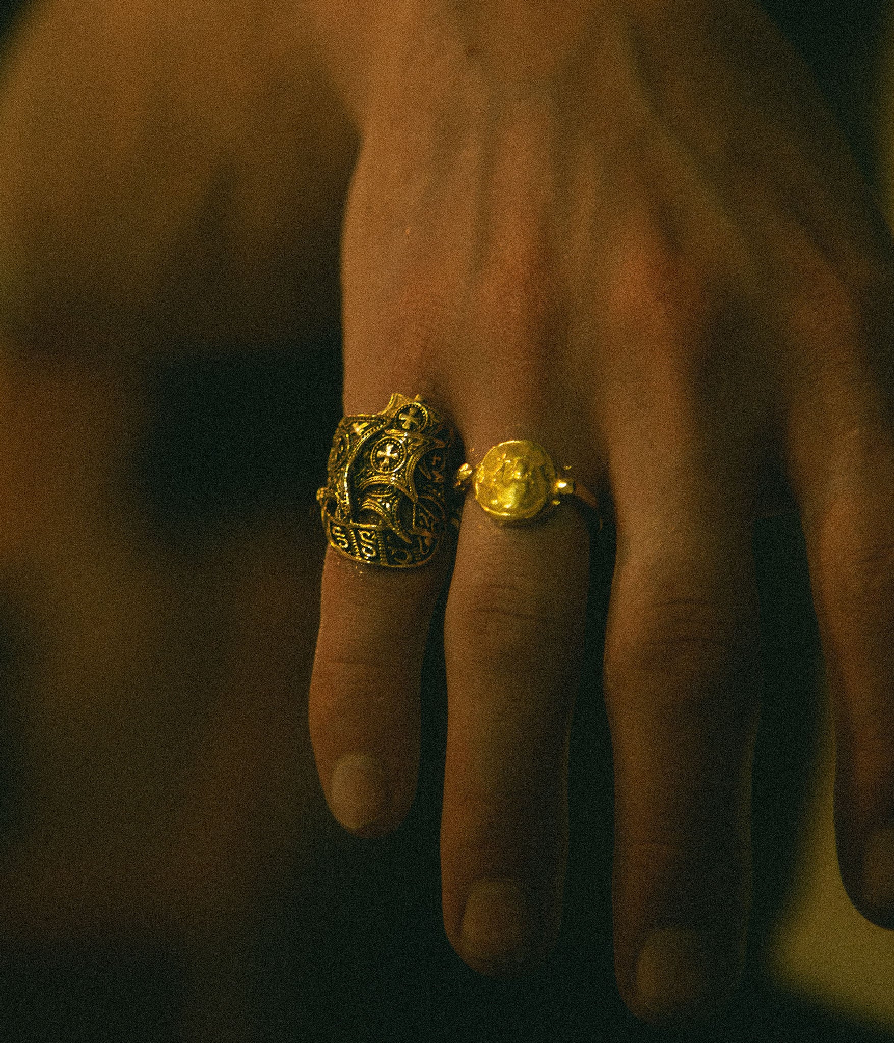 Hand wearing two gold rings with intricate designs on a dark background