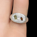 Silver ring with green and brown gemstones on a finger against a black background