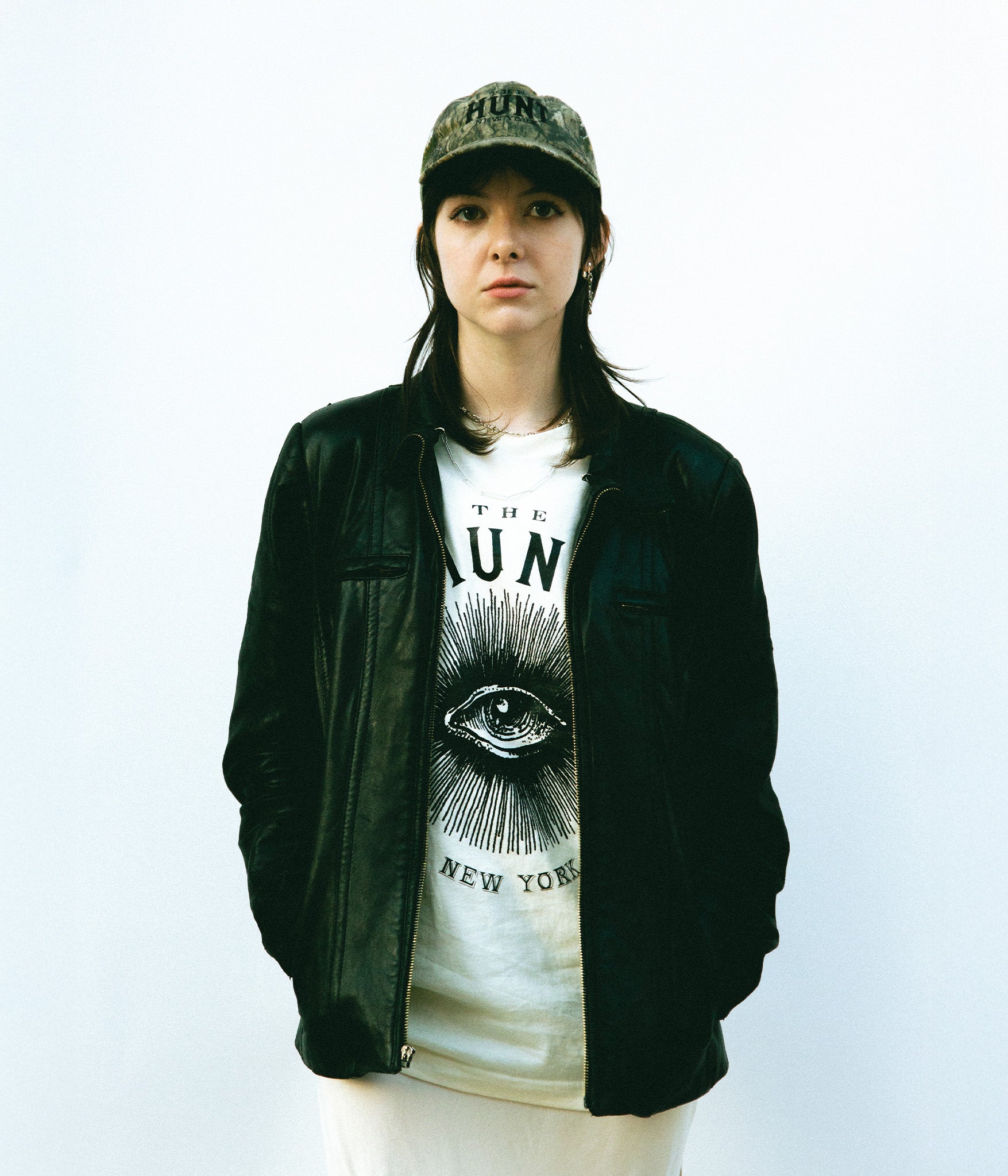 Person wearing a black leather jacket, white t-shirt with graphic design, and camouflage cap on a light gray background