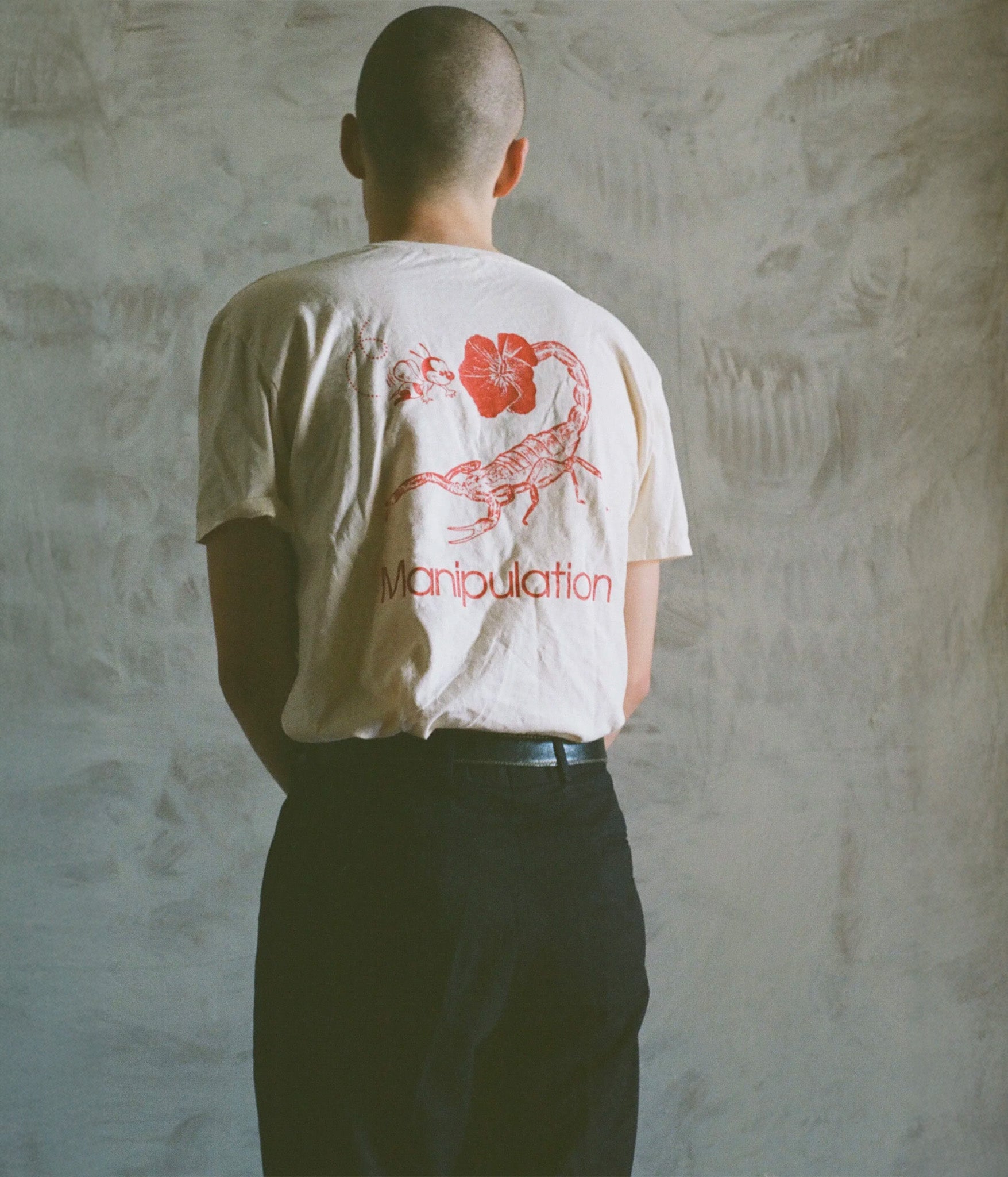 Person wearing a white t-shirt with red graphic and text against a textured wall.