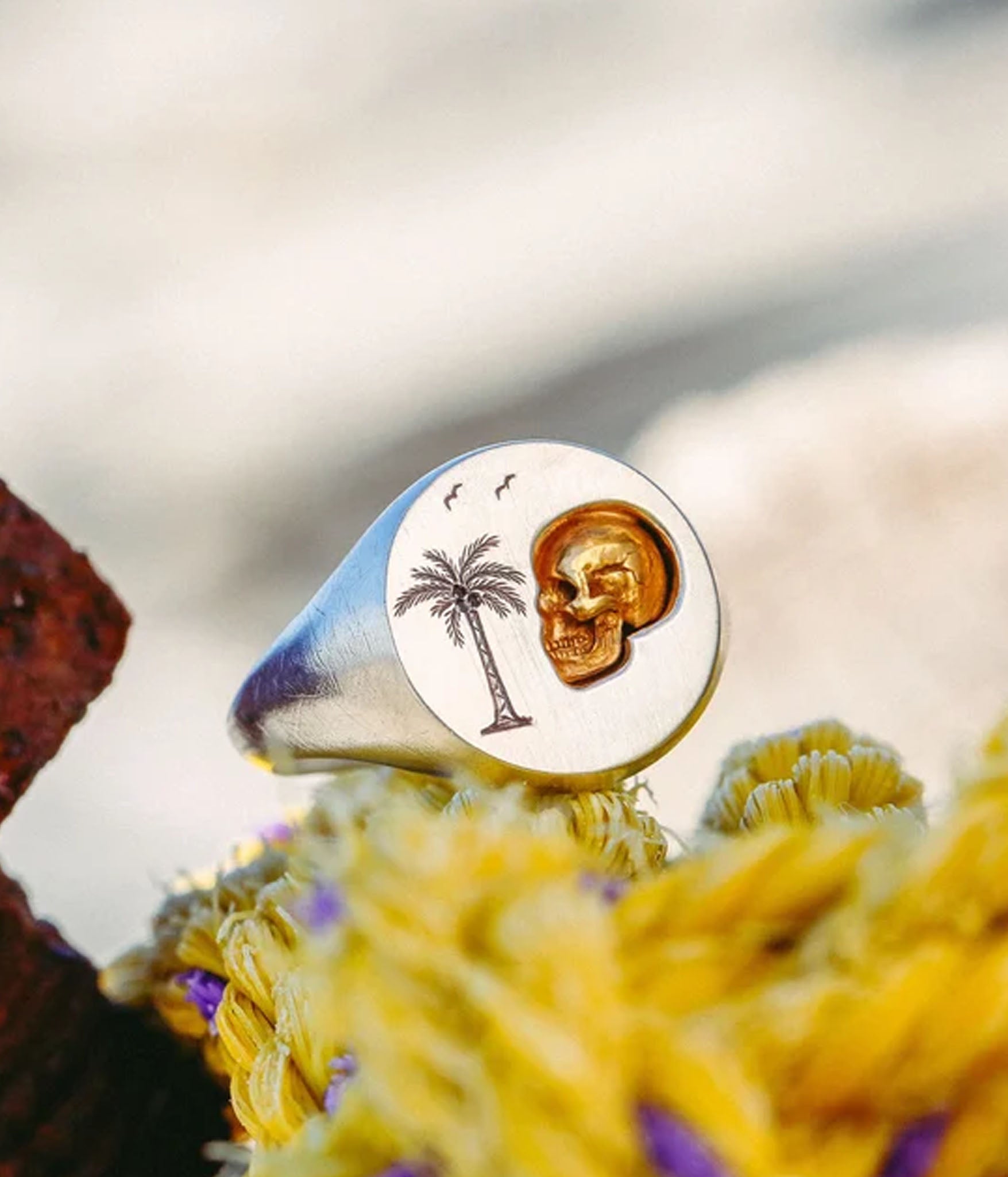 Gold and silver ring with palm tree and skull design on a blurred natural background