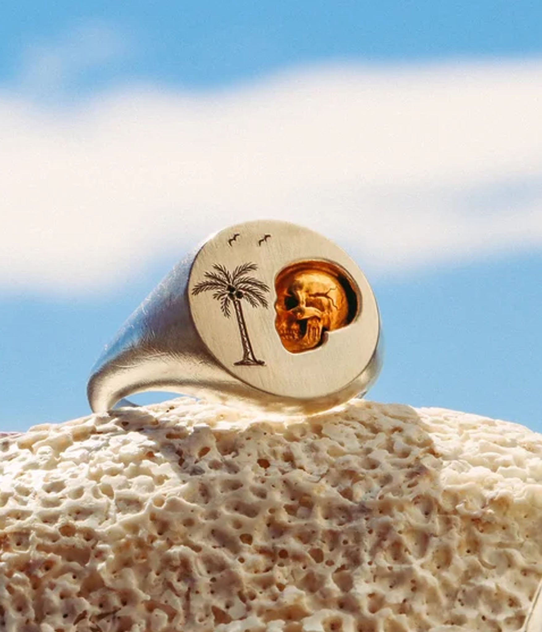 Gold ring with palm tree and skull design on a rock with blue sky background