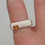 Silver ring with a yellow gemstone on a finger against a neutral background