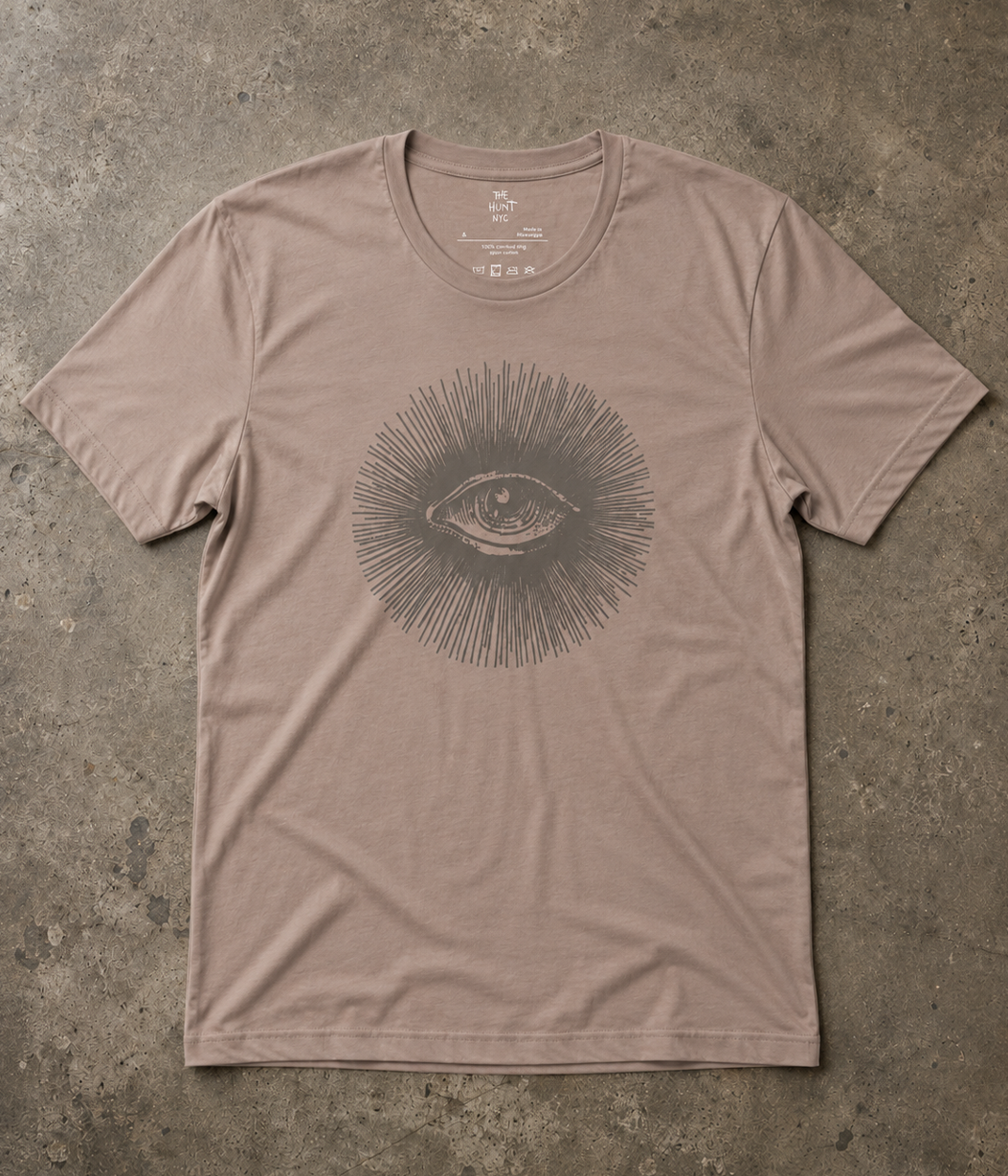 A taupe short-sleeve crewneck t-shirt featuring a dark gray graphic of an eye surrounded by radiating lines, laid flat on a textured concrete surface.