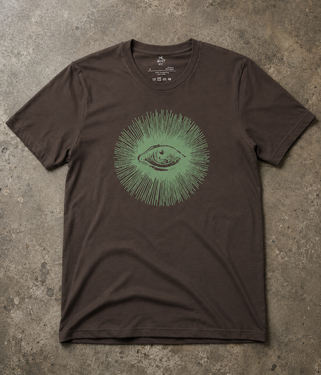 A dark brown short-sleeve t-shirt with a green graphic of an eye surrounded by radiating lines, laid flat on a textured concrete surface.