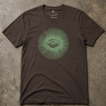 A dark brown short-sleeve t-shirt with a green graphic of an eye surrounded by radiating lines, laid flat on a textured concrete surface.